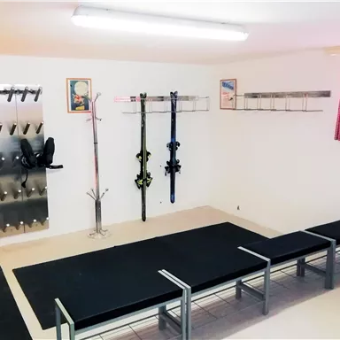 A gear room with shelves for ski equipment and a long area for dressing. The walls are plain, and there is a window with a red curtain.