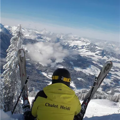 Ein Skifahrer sitzt auf einem verschneiten Hang und blickt auf eine malerische Berglandschaft. Es ist ein klarer blauer Himmel und die Aussicht ist atemberaubend.