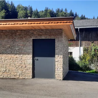 A modern building with a stone facade and a wooden roof. In the background, green trees and another building are visible.