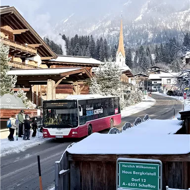A winter view of a picturesque village with snow-covered mountains. A bus stops at a station while passengers wait.