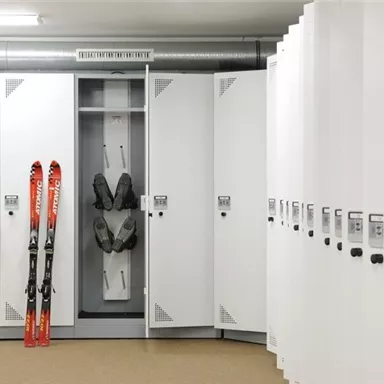 Ein moderner Skiraum mit weißen Schließfächern. Zwei Skier und eine Skiausrüstung sind in einem der Fächer verstaut.