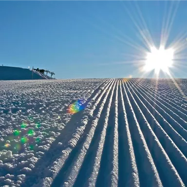 A snow-covered slope with perfectly groomed trails. The sun shines brightly in the clear blue sky.