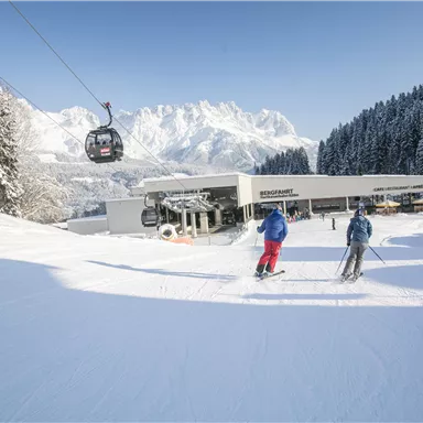 Zwei Skifahrer auf frisch präparierter Piste vor einer Berglandschaft. Im Hintergrund ist eine Kabinenbahn und ein Restaurant zu sehen.