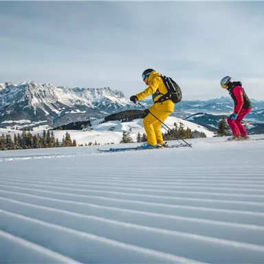 Two skiers are skiing on a freshly prepared slope in the mountains. The landscape is covered in snow, and the mountains are visible in the background.