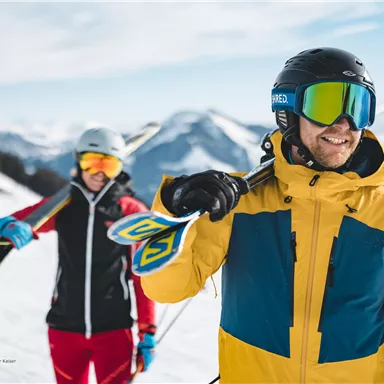 Two skiers are standing on a snow-covered slope in the mountains. The man is wearing a colorful ski suit and is smiling, while the woman in the background looks ready to ski.