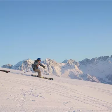 Zwei Skifahrer fahren eine schneebedeckte Piste hinunter. Im Hintergrund sind majestätische Berge zu sehen.
