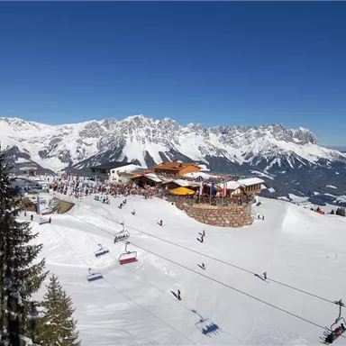 An impressive mountain landscape with snow-covered slopes and a cozy restaurant. Skiers enjoy the sunny day under a clear blue sky.