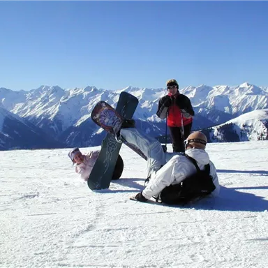 Three people are standing on a snow-covered mountain peak. In the background, there are high mountains and a blue sky.