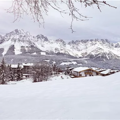 Eine verschneite Landschaft mit majestätischen Bergen im Hintergrund. Im Vordergrund sind einige gemütliche Holzhäuser zu sehen.