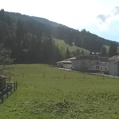 Eine malerische Landschaft mit grünen Wiesen und hügeligen Wäldern. Im Hintergrund befinden sich mehrere Gebäude, die harmonisch in die Natur eingebettet sind.