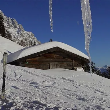 A cozy cabin in the snow with skis in the foreground. The sky is clear and the mountains are visible in the background.