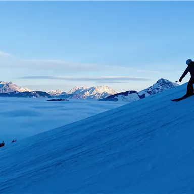 Ein Skifahrer fährt eine verschneite Piste hinunter. Im Hintergrund sind Berge und ein strahlend blauer Himmel zu sehen.