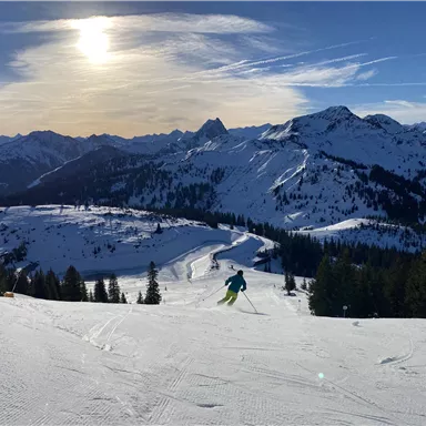 Ein Skifahrer fährt die verschneite Piste hinunter, umgeben von Bergen und einer klaren Winterlandschaft. Die Sonne scheint über den schneebedeckten Gipfeln.
