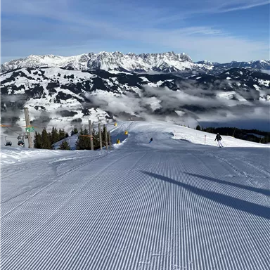 Eine malerische Skipiste mit frisch präpariertem Schnee. Im Hintergrund sind majestätische Berge und ein klarer Himmel zu sehen.