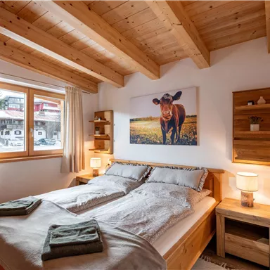 Ein gemütliches Schlafzimmer mit einem Doppelbett und Holzmöbeln. An der Wand hängt ein Bild einer Kuh, und ein Fenster lässt viel Tageslicht herein.