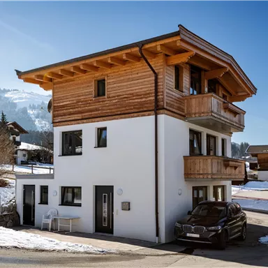 Ein modernes Haus mit einem Holzstuhl und einem Auto vor der Tür. Die Umgebung ist schneebedeckt und die Berge sind im Hintergrund sichtbar.