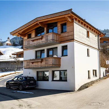 Ein modernes, zweigeschossiges Haus mit Holzbalustraden in einer schneebedeckten Landschaft. Im Vordergrund steht ein Auto und im Hintergrund sind Berge zu sehen.