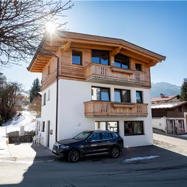 Ein modernes Haus mit Holzelementen und großen Fenstern. Schnee liegt auf dem Boden und die Berge sind im Hintergrund sichtbar.