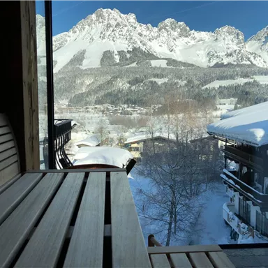 Ein Blick aus einem Fenster auf beeindruckende schneebedeckte Berge. Im Vordergrund ist eine Holzbänke zu sehen, die eine gemütliche Atmosphäre vermittelt.