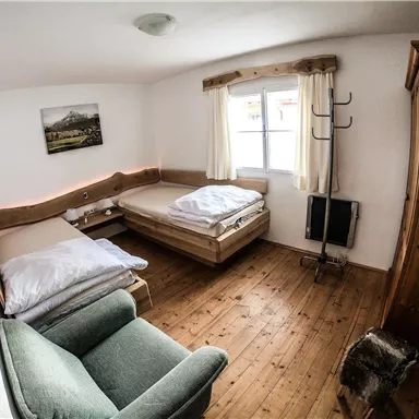 A simple room with two beds and a cozy armchair. The wooden floor and the bright windows create an inviting atmosphere.
