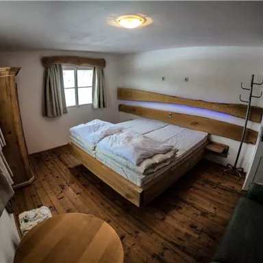 A simple bedroom with a double bed and wooden furniture. The room has a window with curtains and a small table.