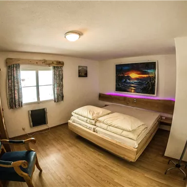 A cozy bedroom with a double bed and a blue armchair. A colorful picture of a sunset hangs on the wall.