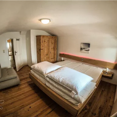 A cozy bedroom with a large bed and wooden floor. There is a wardrobe and a small seating area.