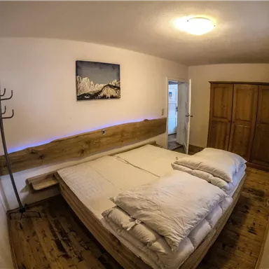 A cozy bedroom with a large bed and bright pillows. On the wall hangs a picture of mountains, and there is a wooden wardrobe as well as a coat rack.