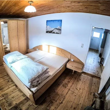 A cozy bedroom with a wooden bed and large pillows. The walls are painted light and the wooden ceiling gives a warm impression.