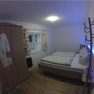 A simple bedroom with a double bed and a wardrobe. The walls are light, and there is a window with curtains.