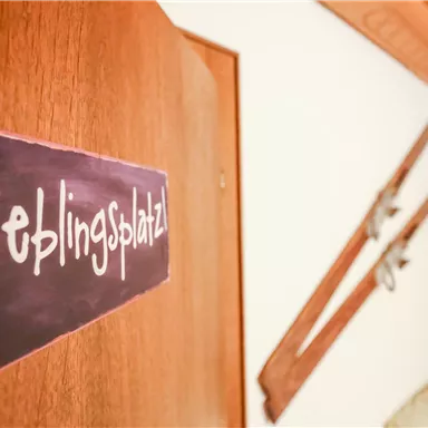 Eine Tür mit einem Schild, das "Lieblingsplatz" zeigt. Im Hintergrund sind Skier und ein gemütlicher Raum zu sehen.