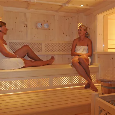 Zwei Frauen entspannen in einer hellen Holzsauna. Die Umgebung ist gemütlich und einladend.