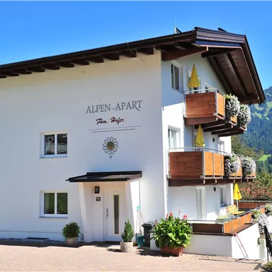 A modern white building named "Alpen-Apart" with charming balconies. In the background, there are green mountains and a clear blue sky.
