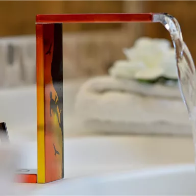 A modern faucet with a colorful design from which water flows. In the background, white towels and a flower are visible.