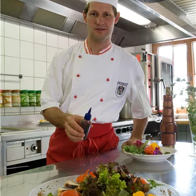 A chef is standing in a modern kitchen and showcasing a colorful salad on a plate. In the background, kitchen utensils and fresh ingredients are visible.