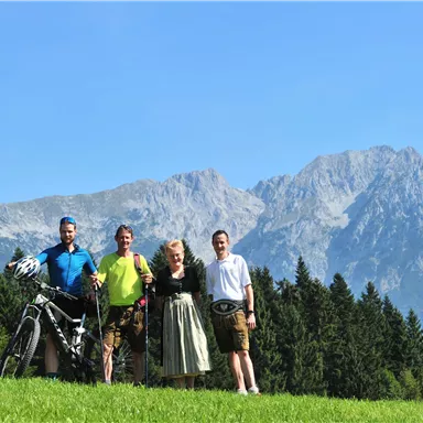 A group of four people stands on a meadow with mountains in the background. The men are wearing sporty clothing and the woman is wearing a traditional dress.