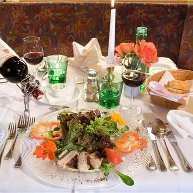 An elegantly set table with a colorful salad, grilled meat, and a bottle of red wine. There are glasses and fresh flowers in an appealing setting.