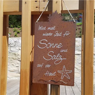 A wooden sign hangs on a wooden railing. It reads: "It's time again for sun and salt on the skin."