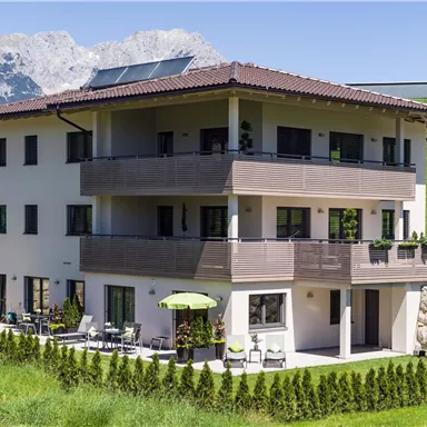 A modern building with several floors and balconies. In the background, mountains and a green hillside are visible.