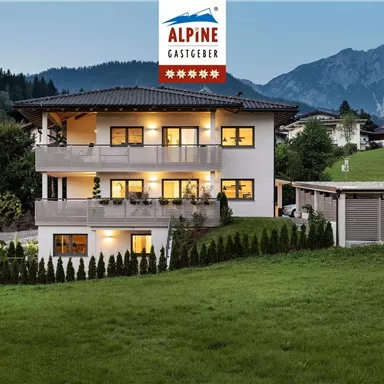 A modern house with several floors in an idyllic setting. In the background, mountains and a clear sky can be seen.