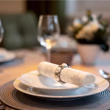 An elegantly set table with a white plate and a decorative napkin. In the background, wine glasses and a floral arrangement can be seen.