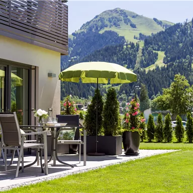A beautiful outdoor area with a table and chairs, as well as a sunny sun umbrella. In the background, green mountains and a well-maintained lawn can be seen.