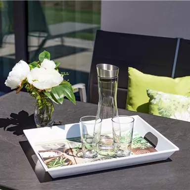 An outdoor table with a white serving platter, on which a glass carafe and glasses are placed. Next to it is a vase with flowers and a few decorative pillows.