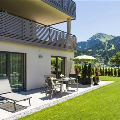 A modern terrace with garden furniture and a parasol. In the background, green hills and a clear blue sky can be seen.