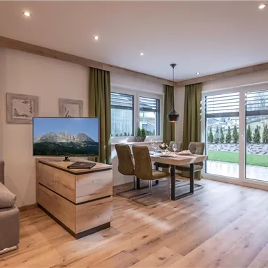 A modern living room with a cozy sofa and a television. The dining area has a table with chairs and large windows that look out onto a garden.