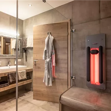 A modern bathroom with concrete walls and an elegant wooden countertop. There is a towel rack, a mirror, and a red infrared light source on the wall.