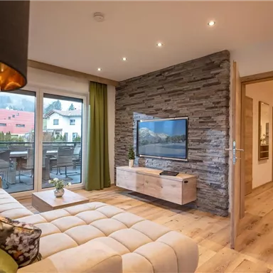 A modern living room with a comfortable sofa and a television. Large windows offer a view of a balcony and plenty of natural light.