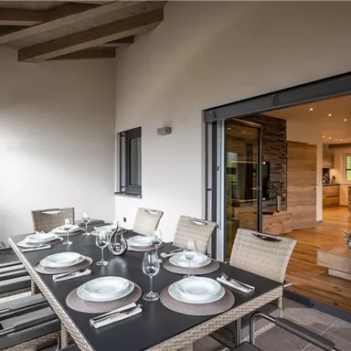 A stylish outdoor dining area with a large table and elegant chairs. In the background, a modern kitchen is visible.