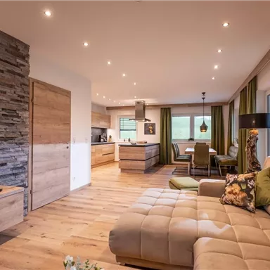 A modern living room with a comfortable sofa and a wooden floor. In the background, there is an open kitchen and a dining area with large windows.