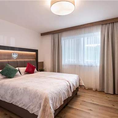 A modern bedroom with two beds, a large window, and wooden-colored furnishings. The pillows are designed in various colors, creating a cozy atmosphere.
